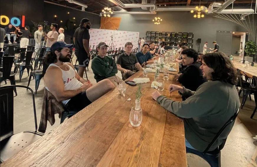 A long wooden table of community builders sits deep in conversation at a Skool meetup, with Matt seated among them halfway down the table.